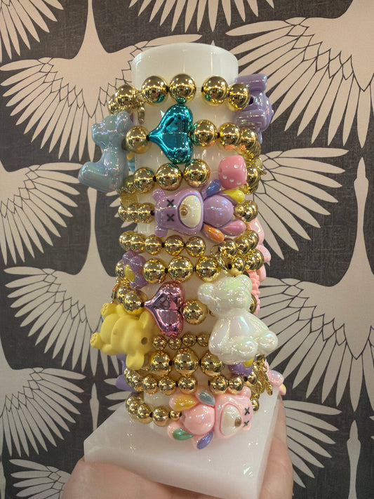 Adorable girls’ elastic bracelets with chunky gold beads and iridescent pastel bear, heart, flower, dice and gold bike charms. Perfect for stacking!