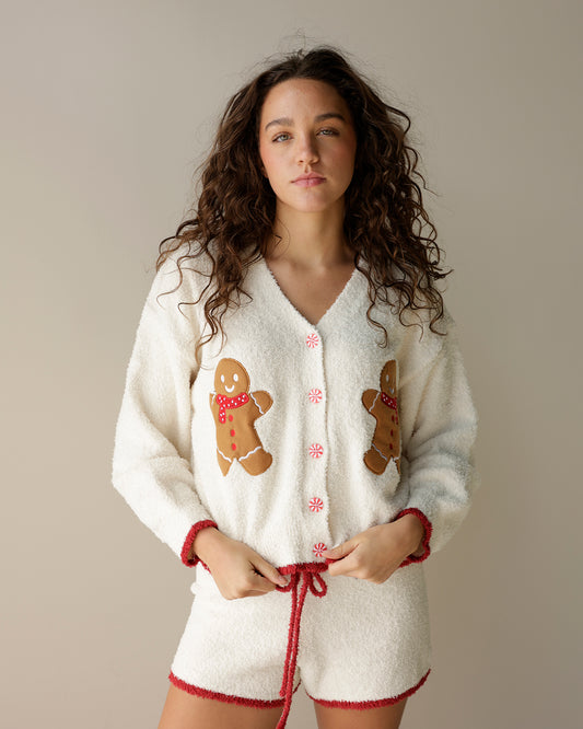 A woman wearing a cozy white bouclé pajama set with gingerbread men applique and red trim, standing against a plain beige studio background.