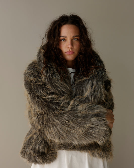 A woman with long, wavy dark brown hair is wearing a voluminous, shaggy faux fur wrap or jacket in a mottled gray, brown, and black pattern, styled over a white top against a plain, warm gray studio background.
