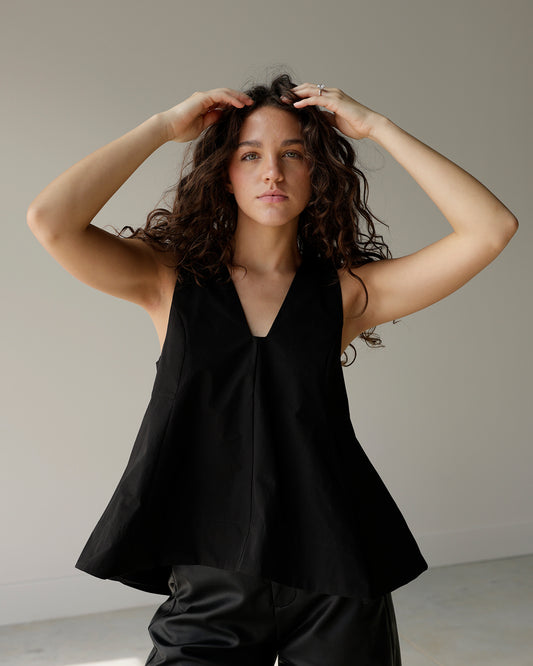 woman wearing a black sleeveless maude top with a deep v-neckline and dramatic flared a-line hem, styled with black faux leather pants.