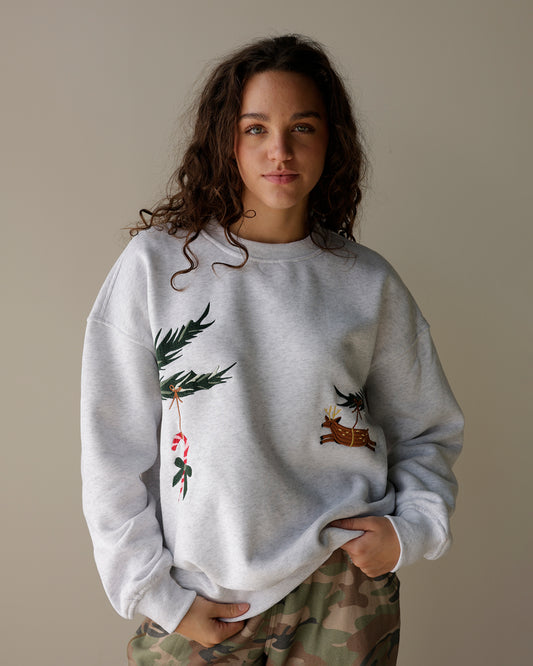 woman wearing a light heather grey le lis christmas sweatshirt with embroidered evergreen branches, candy cane and reindeer, styled with camo cargo pants.
