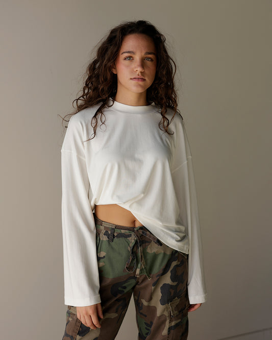 A woman with long, wavy brown hair stands against a plain beige studio background, wearing a white long-sleeve crop top and green camouflage drawstring pants.