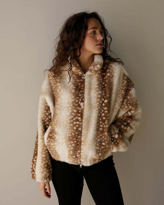 A woman wearing a brown and cream faux fur jacket with a white spotted deer-antler pattern, paired with black pants, standing against a plain beige background.