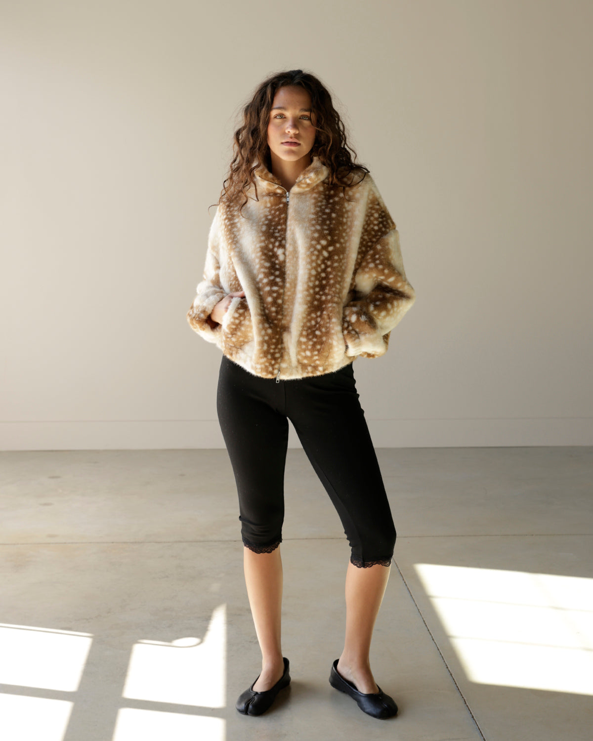 A woman stands in a minimalist studio wearing a fuzzy beige and brown spotted jacket, black cropped leggings, and black flats.