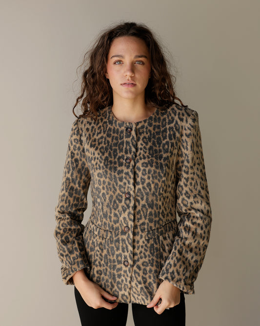 A woman modeling a leopard print button-up jacket with structured shoulders and front pockets, set against a plain warm gray studio background.