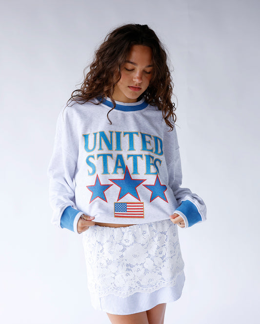 United States sweatshirt featuring blue outlined gold lettering, red-outlined stars, American flag detail, and blue-gray trim. A cozy patriotic pullover perfect for everyday wear and holidays.
