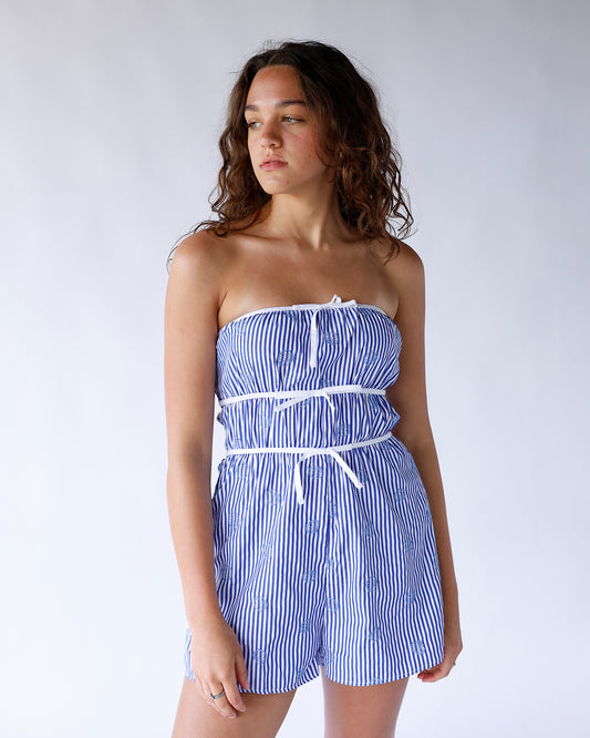 Blue and white striped strapless romper featuring blue seashell embroidery and three vertical white bows. A cute coastal-inspired one-piece perfect for summer and vacation wear.