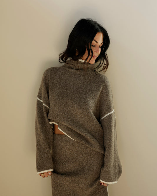 A person's hand holding up a gray wool turtleneck poncho sweater with white contrast stitching, photographed in a retail store setting.