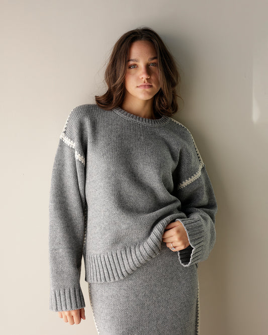 A close-up of a gray knit sweater hanging on a wooden hanger, showing the ribbed crew neck and a white decorative zigzag stitch detail along the sleeve.