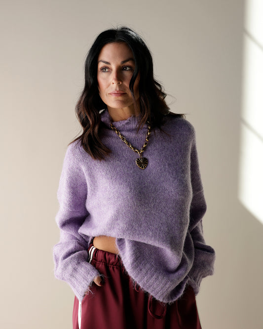 A lavender purple knit mock-neck sweater hanging on a wooden hanger, with other garments partially visible on the sides.