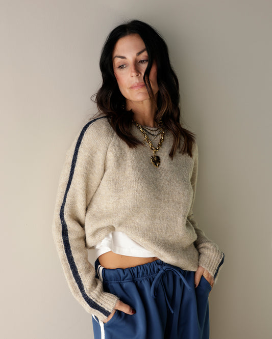 A fuzzy knit crewneck sweater in light oatmeal beige hanging on a wooden hanger, with navy and brown striped sweaters visible behind it on a clothing rack.