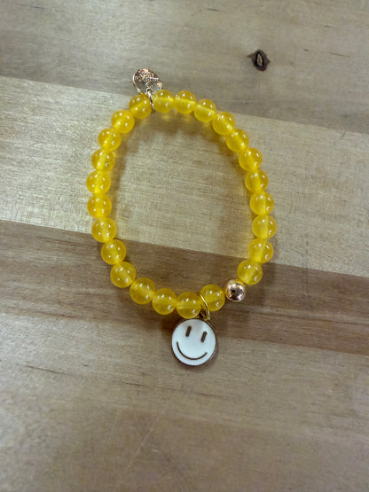 Yellow beaded stretch bracelet with white enamel smiley face charm on wood surface