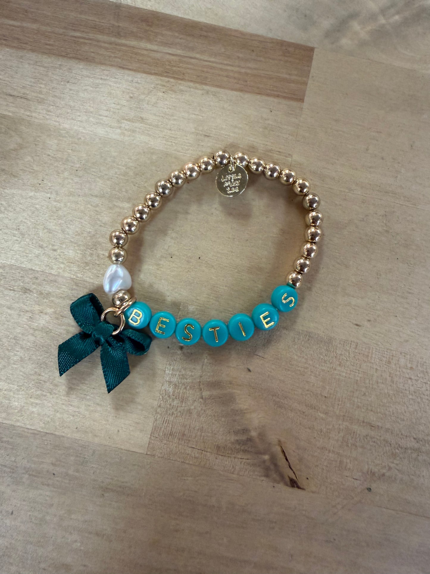 Gold beaded stretch bracelet with teal letter beads spelling BESTIES, bow charm, and pearl