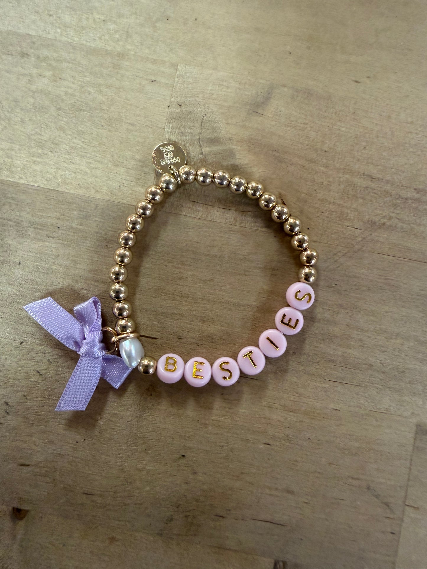 Gold beaded bracelet with pink letter beads spelling BESTIES and lavender bow charm