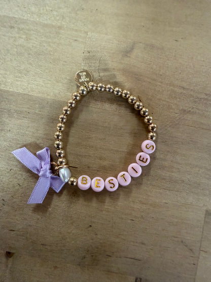 Gold beaded bracelet with pink letter beads spelling BESTIES and lavender bow charm