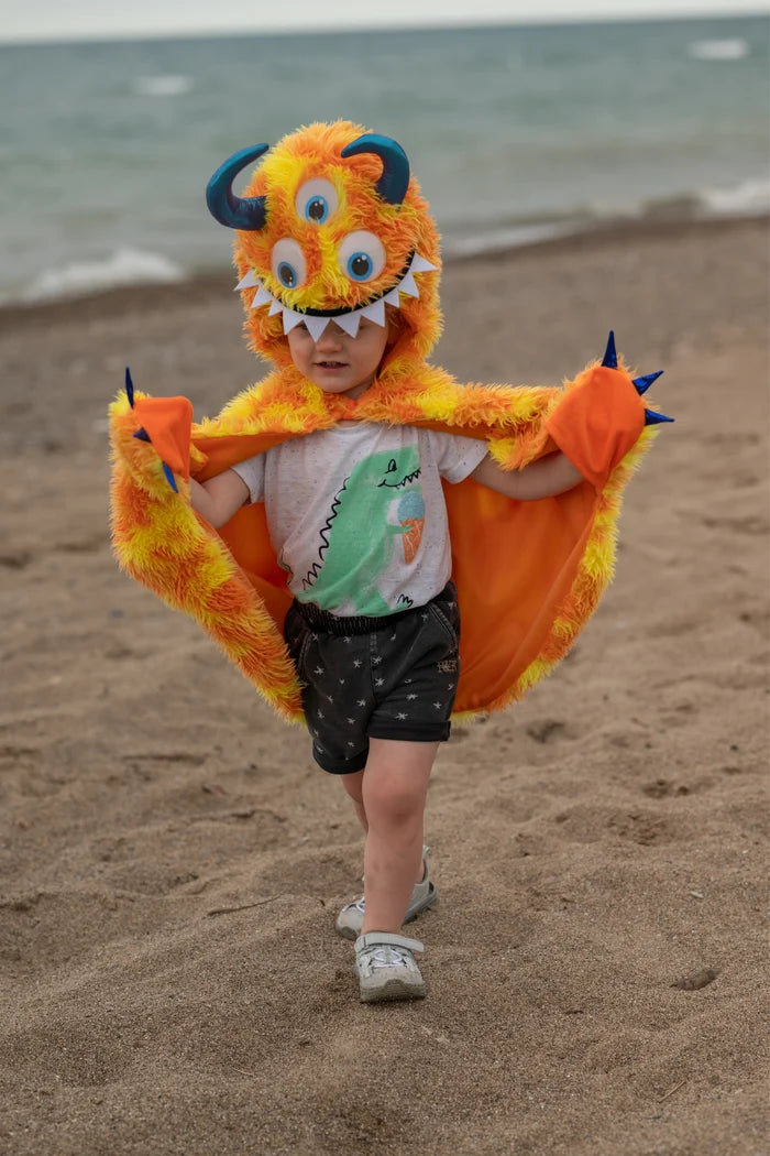 Child wearing orange fuzzy monster cape with hood, horns, and claws at beach
