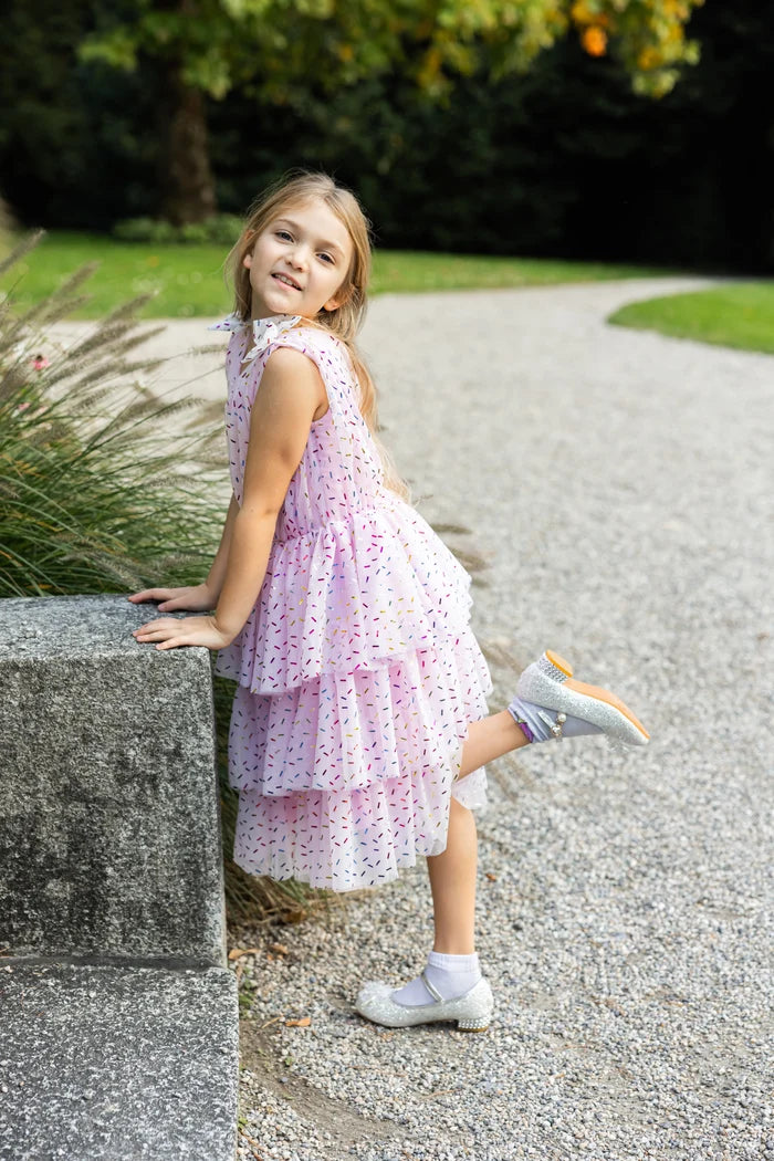 Girl wearing pink tiered ruffle dress with colorful sprinkle print and white shoes
