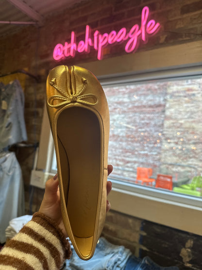 Gold metallic split-toe ballet flat with bow detail, held up in store