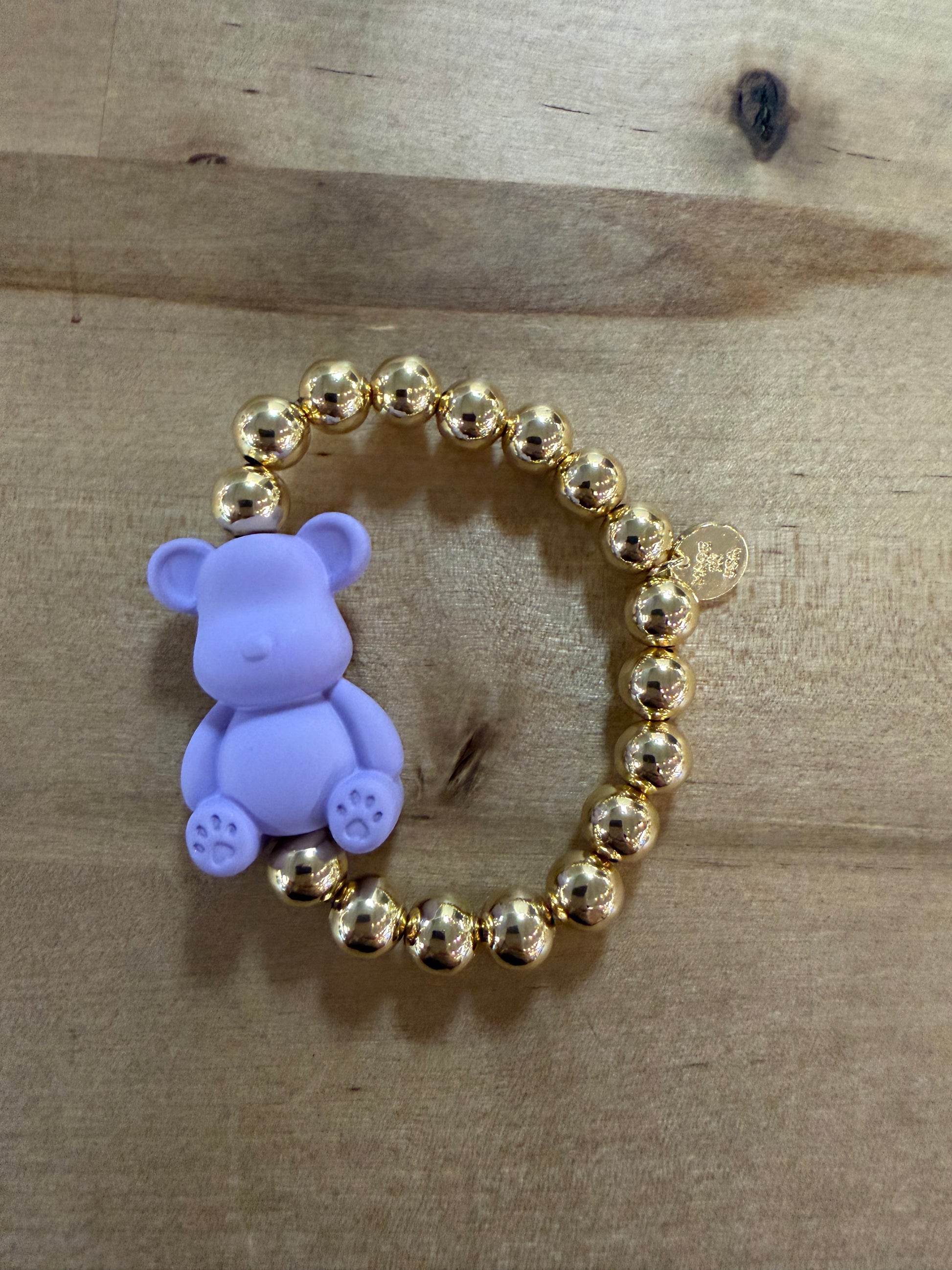 Gold beaded stretch bracelet with lavender silicone teddy bear charm on wood surface