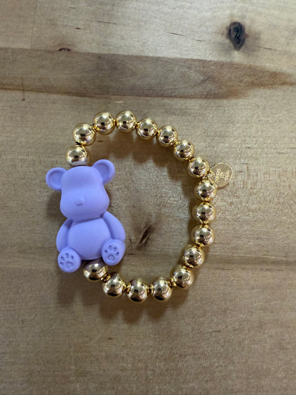 Gold beaded stretch bracelet with lavender silicone teddy bear charm on wood surface