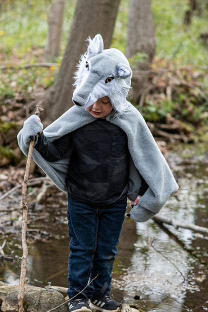 Child wearing gray wolf cape costume with hood, holding stick in forest