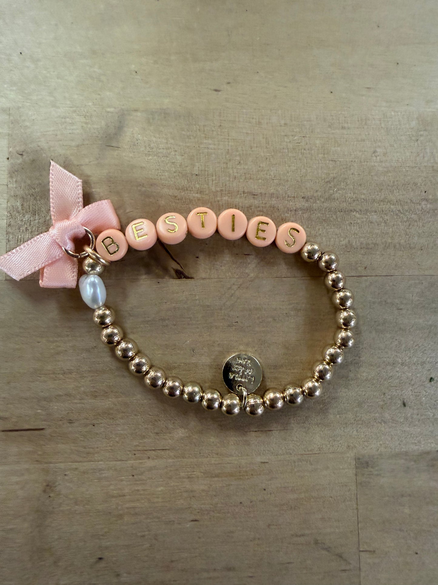 Gold beaded bracelet with pink letter beads spelling BESTIES, pink bow, and pearl charm