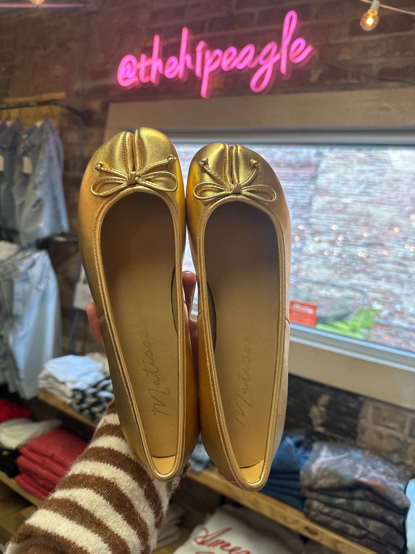 Gold metallic split-toe ballet flats with bow detail, held up in store