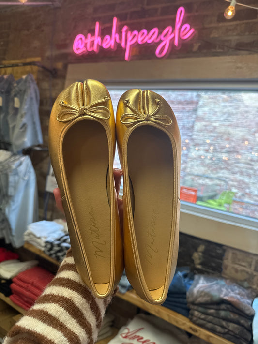 Gold metallic split-toe ballet flats with bow detail, held up in store