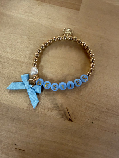 Gold beaded bracelet spelling BESTIES in blue letter beads with a light blue bow charm
