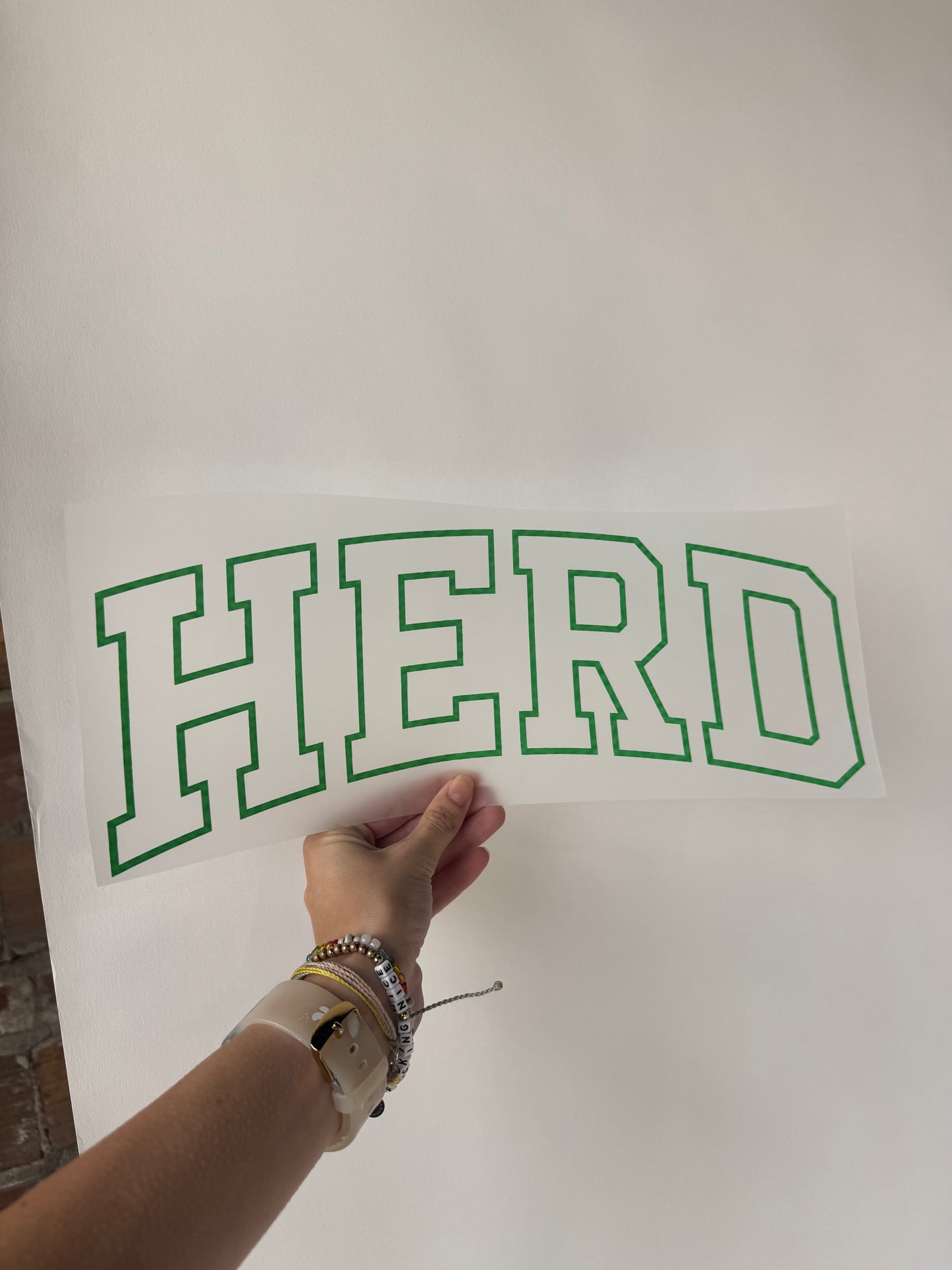 Hand holding white decal with green outlined block letters spelling HERD