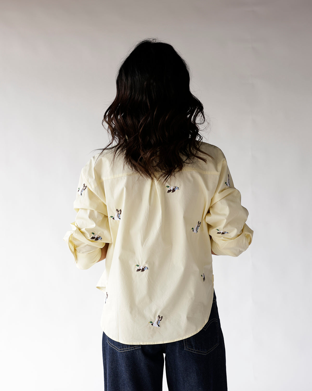 Cream button-up shirt with duck embroidery, worn from the back, paired with dark jeans.