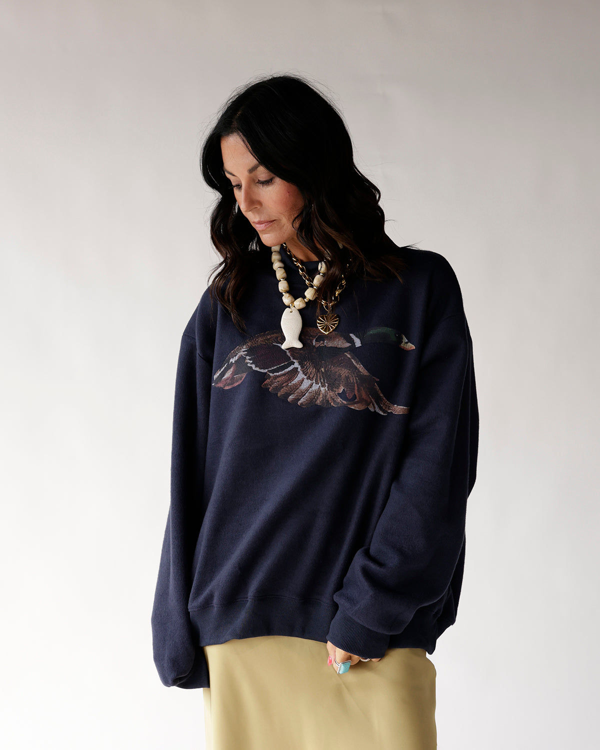 Oversized navy sweatshirt with mallard duck graphic, worn with yellow skirt and layered necklaces.