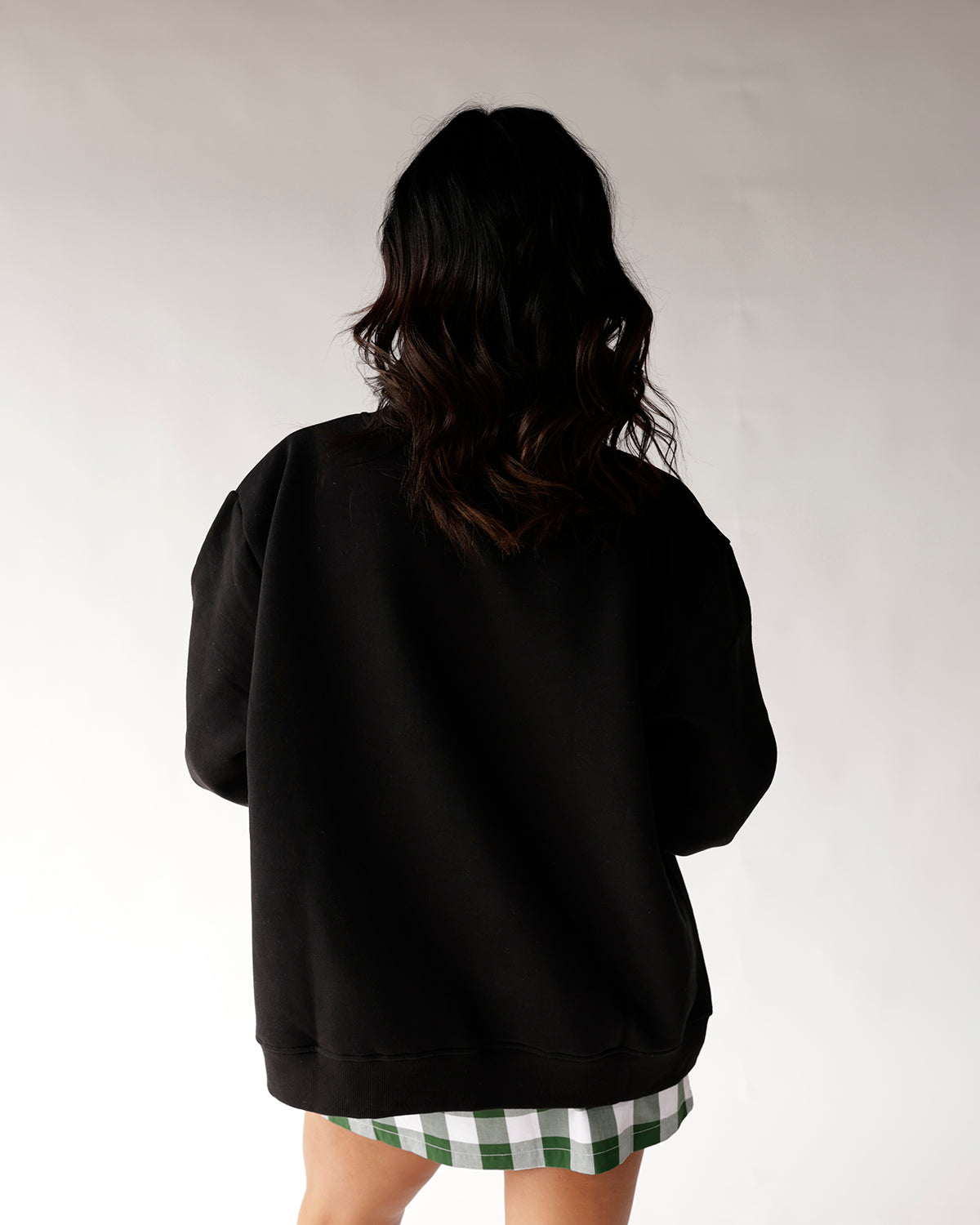 Oversized black mock neck sweatshirt shown from back, paired with green plaid skirt.