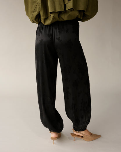 Black satin jacquard floral jogger pants with elastic cuffs, worn with nude heels