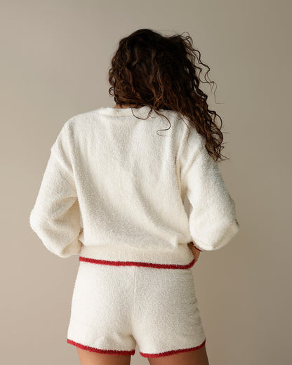 A woman viewed from behind wearing a cozy, textured off-white fleece loungewear set with a matching sweatshirt and shorts, featuring a thin red trim detail at the waist and hem.