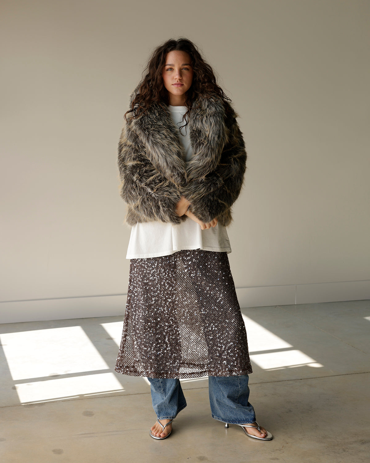 A model stands in a minimalist studio setting wearing a shaggy faux fur jacket, a white top, a sheer patterned skirt, and blue jeans.