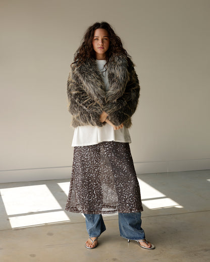 A model stands in a minimalist studio setting wearing a shaggy faux fur jacket, a white top, a sheer patterned skirt, and blue jeans.