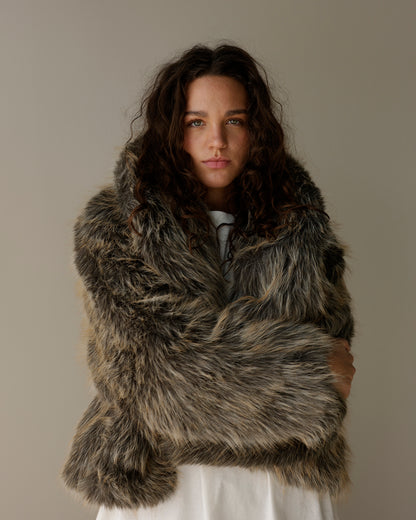 A woman with long, wavy dark brown hair is wearing a voluminous, shaggy faux fur wrap or jacket in a mottled gray, brown, and black pattern, styled over a white top against a plain, warm gray studio background.