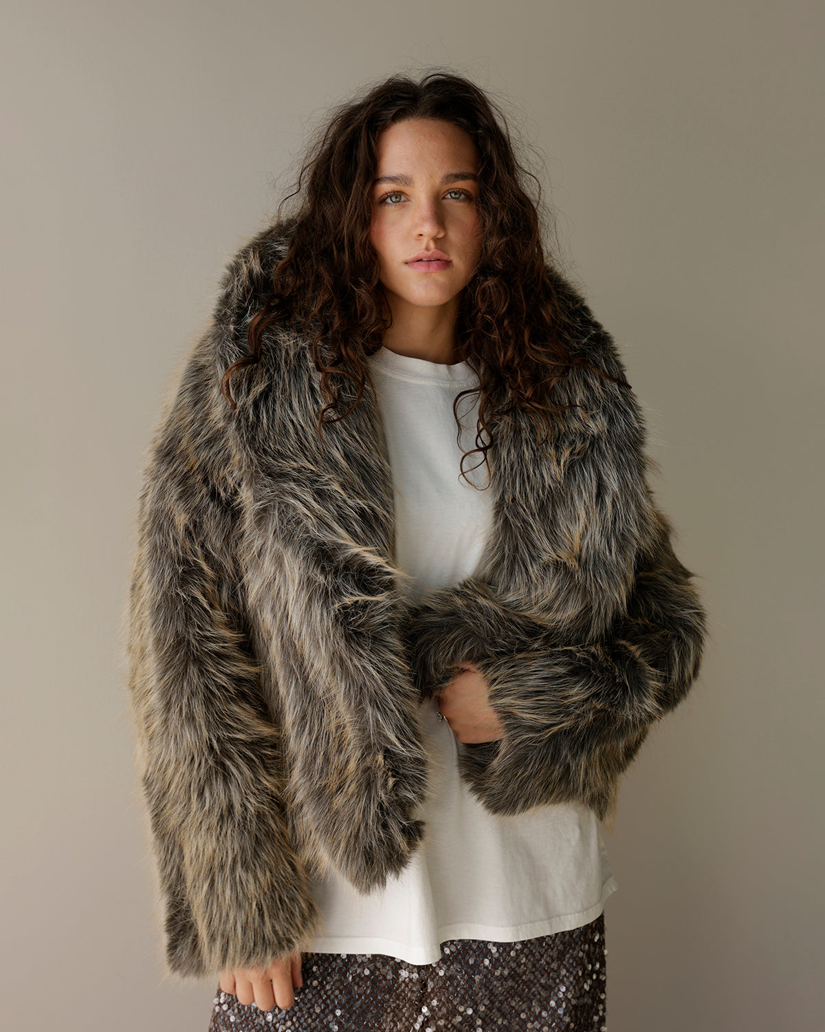 A woman with long, curly brown hair models a shaggy faux fur jacket in a mottled gray, brown, and black pattern, worn over a white t-shirt and a sparkly dark skirt, against a plain beige studio background.