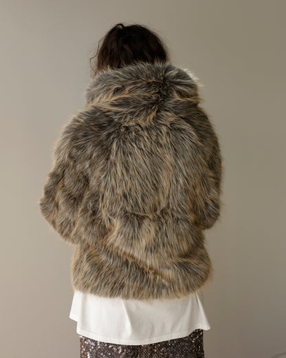 A person viewed from behind wearing a shaggy faux-fur hooded jacket in mixed brown, gray, and beige tones, over a white top and patterned bottoms, against a plain light gray wall.