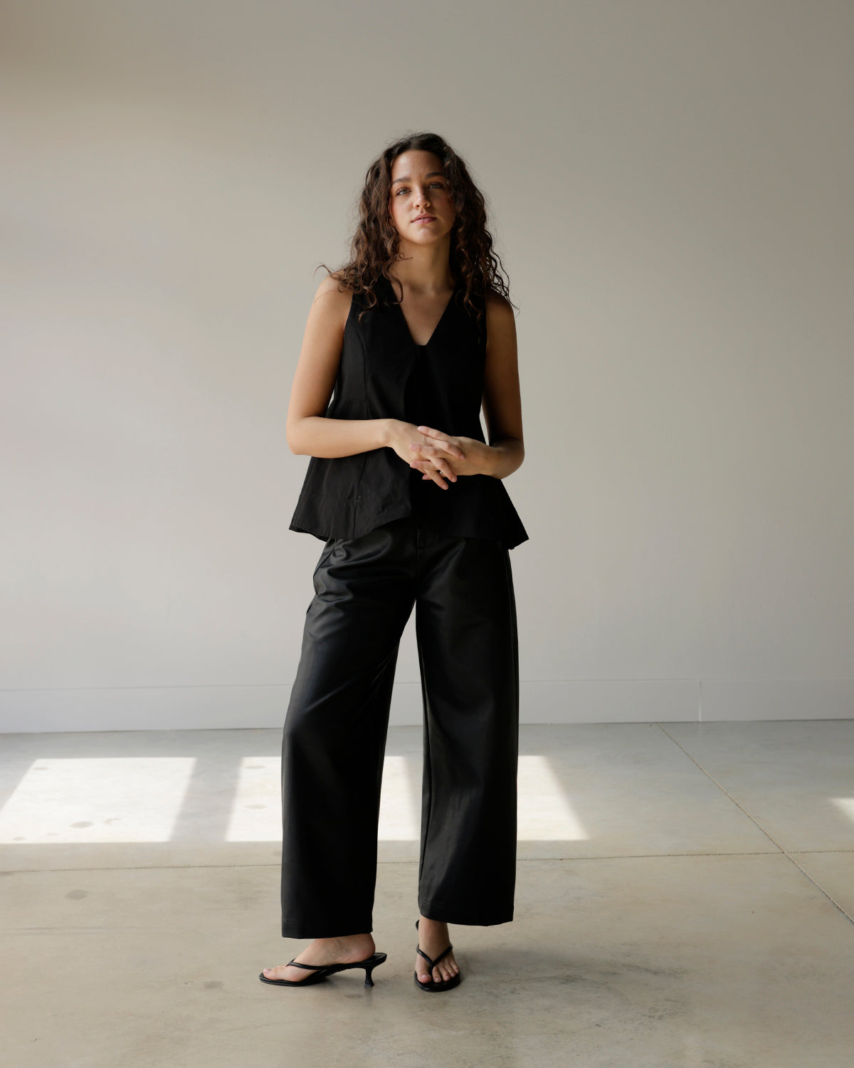 woman wearing a black sleeveless maude top with a deep v-neckline and dramatic flared a-line hem, styled with black faux leather pants.