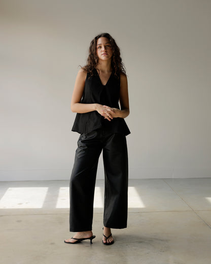 woman wearing a black sleeveless maude top with a deep v-neckline and dramatic flared a-line hem, styled with black faux leather pants.