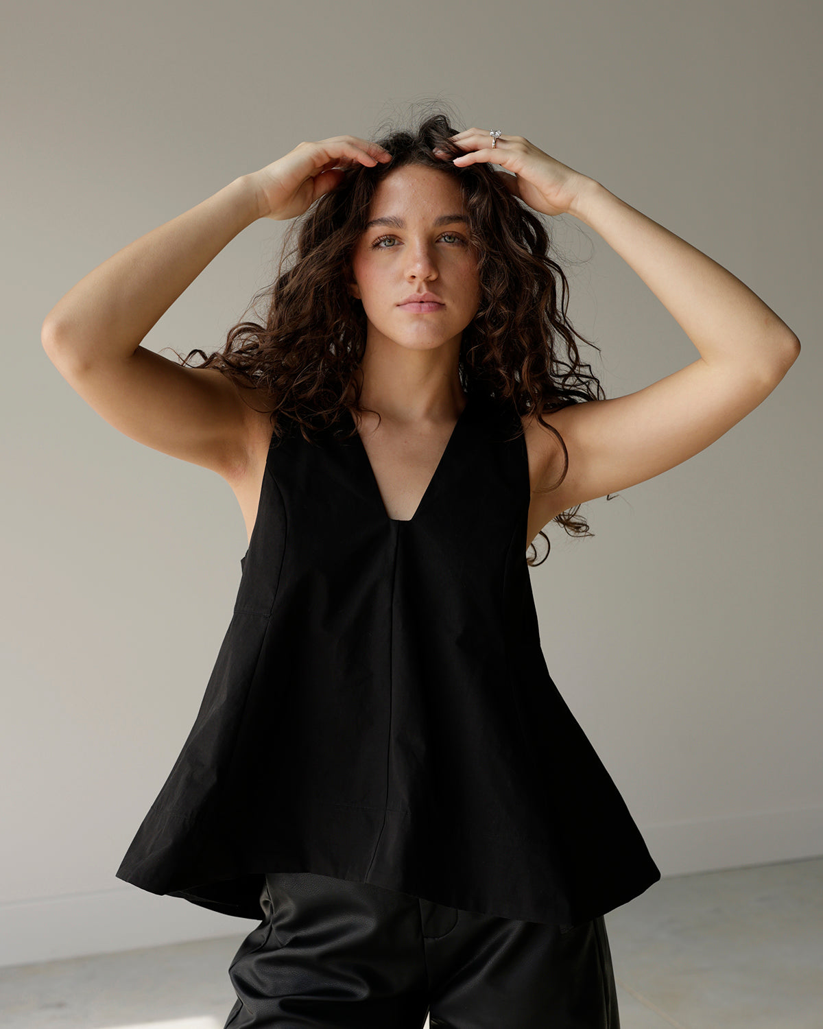 woman wearing a black sleeveless maude top with a deep v-neckline and dramatic flared a-line hem, styled with black faux leather pants.