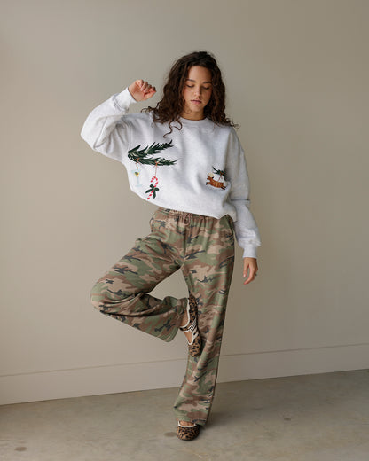 woman wearing a light heather grey le lis christmas sweatshirt with embroidered evergreen branches, candy cane and reindeer, styled with camo cargo pants.