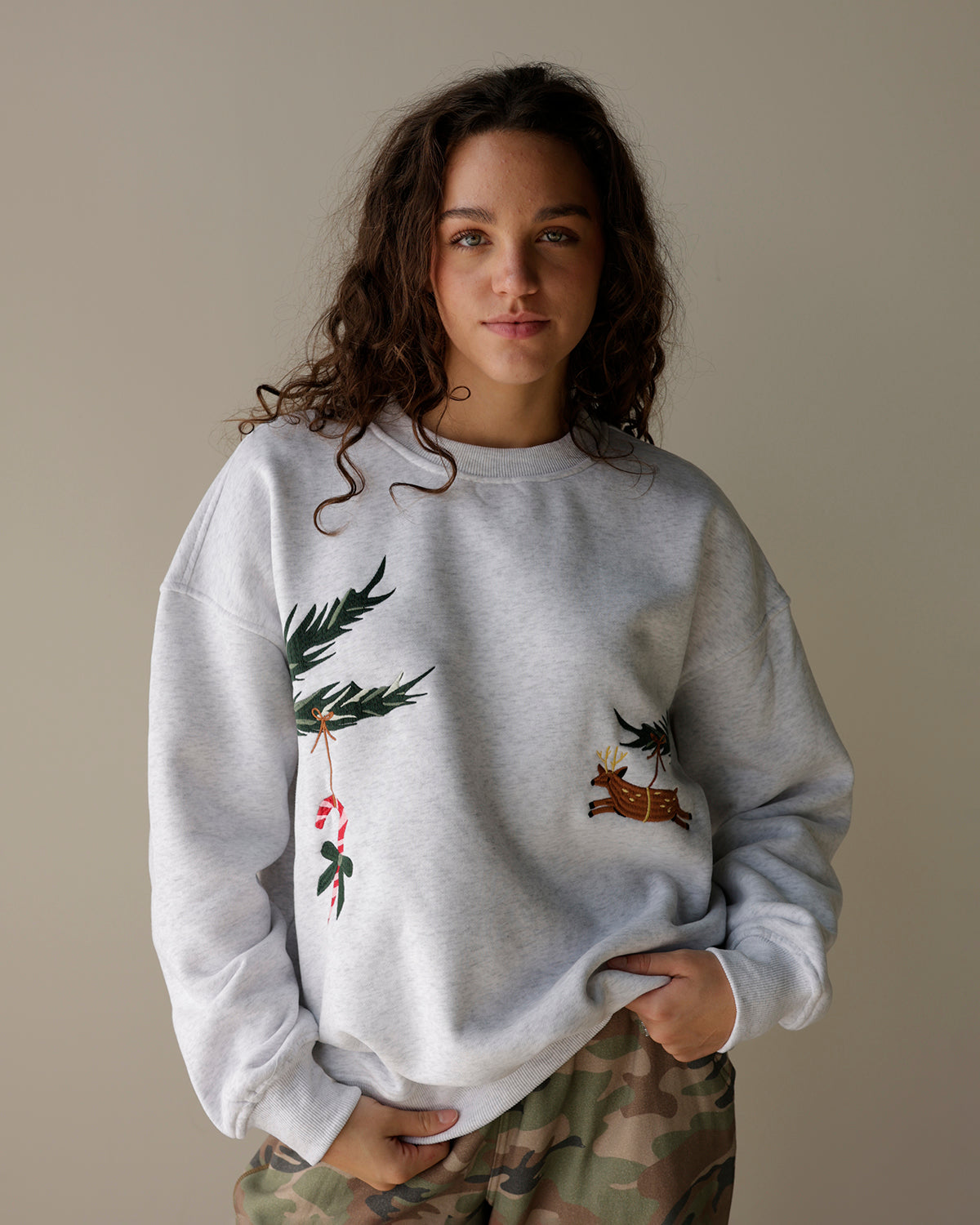 woman wearing a light heather grey le lis christmas sweatshirt with embroidered evergreen branches, candy cane and reindeer, styled with camo cargo pants.