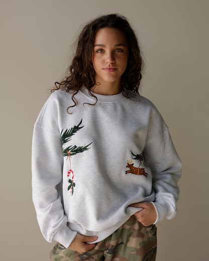 woman wearing a light heather grey le lis christmas sweatshirt with embroidered evergreen branches, candy cane and reindeer, styled with camo cargo pants.
