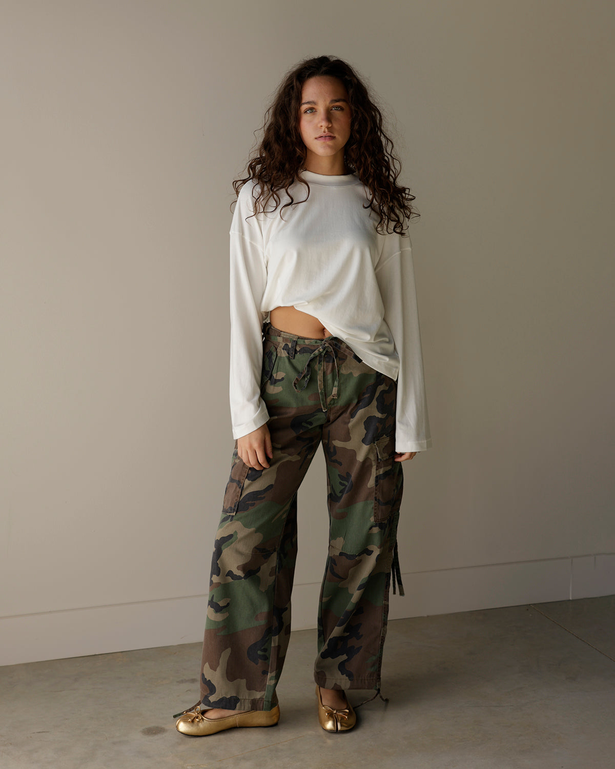 A woman stands in a studio wearing a white cropped long-sleeve top and loose camouflage print pants with metallic gold shoes, against a plain beige wall and concrete floor.