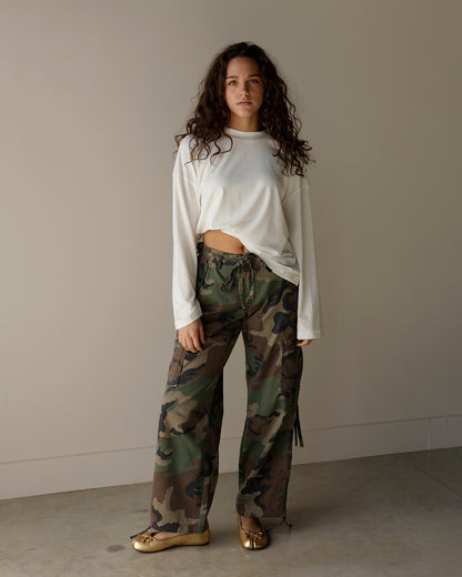 A woman stands in a studio wearing a white cropped long-sleeve top and loose camouflage print pants with metallic gold shoes, against a plain beige wall and concrete floor.