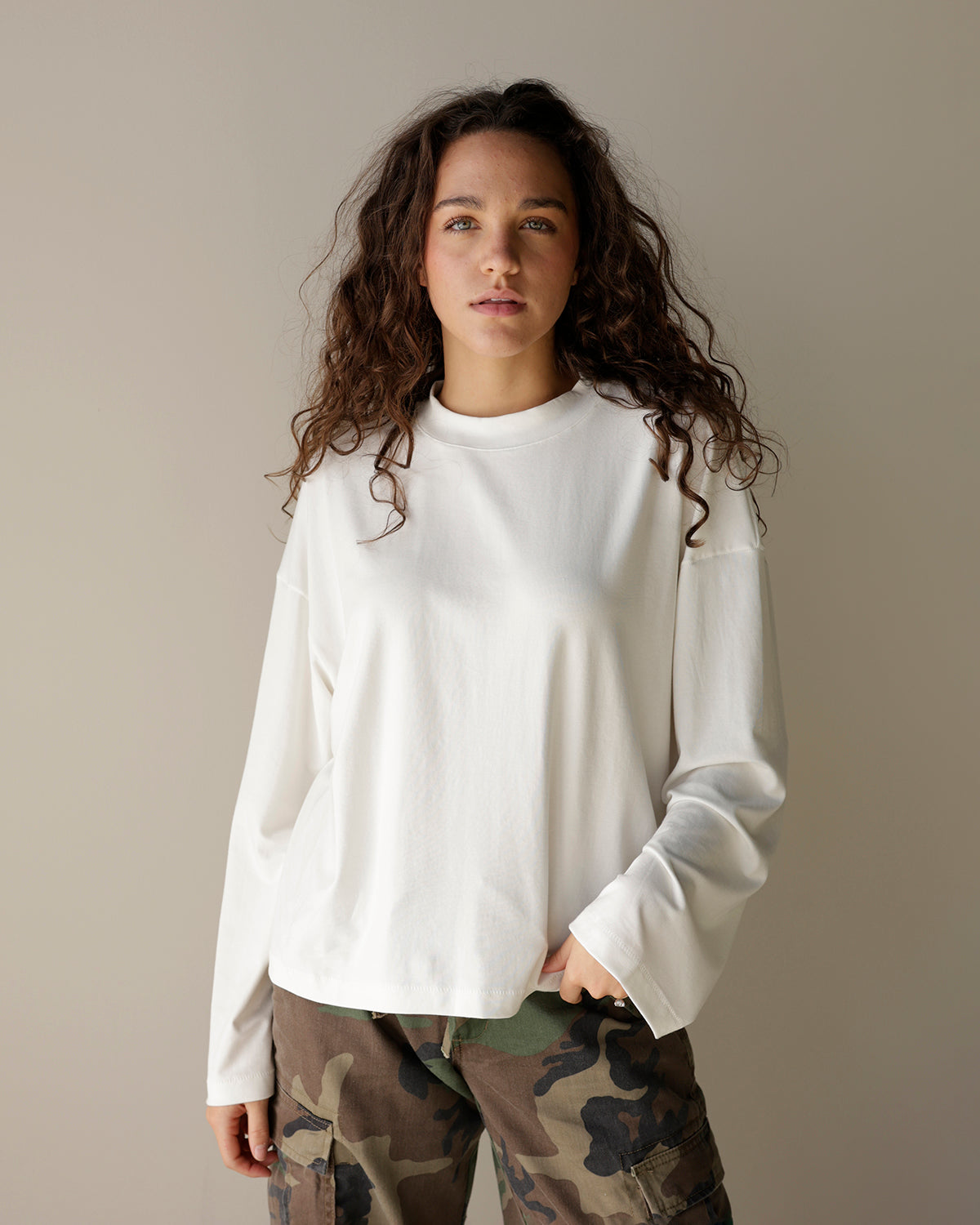 A woman with long, curly brown hair wearing a white long-sleeve crewneck top and camouflage cargo pants, standing against a plain beige studio background.