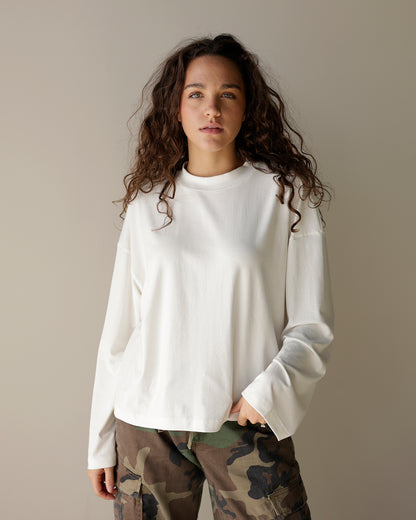 A woman with long, curly brown hair wearing a white long-sleeve crewneck top and camouflage cargo pants, standing against a plain beige studio background.